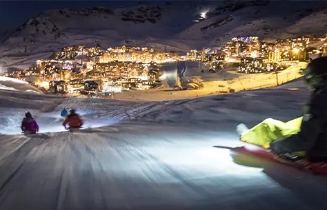 Des personnes font une descente de piste de ski en luge la nuit avec en fond la station de ski illuminée